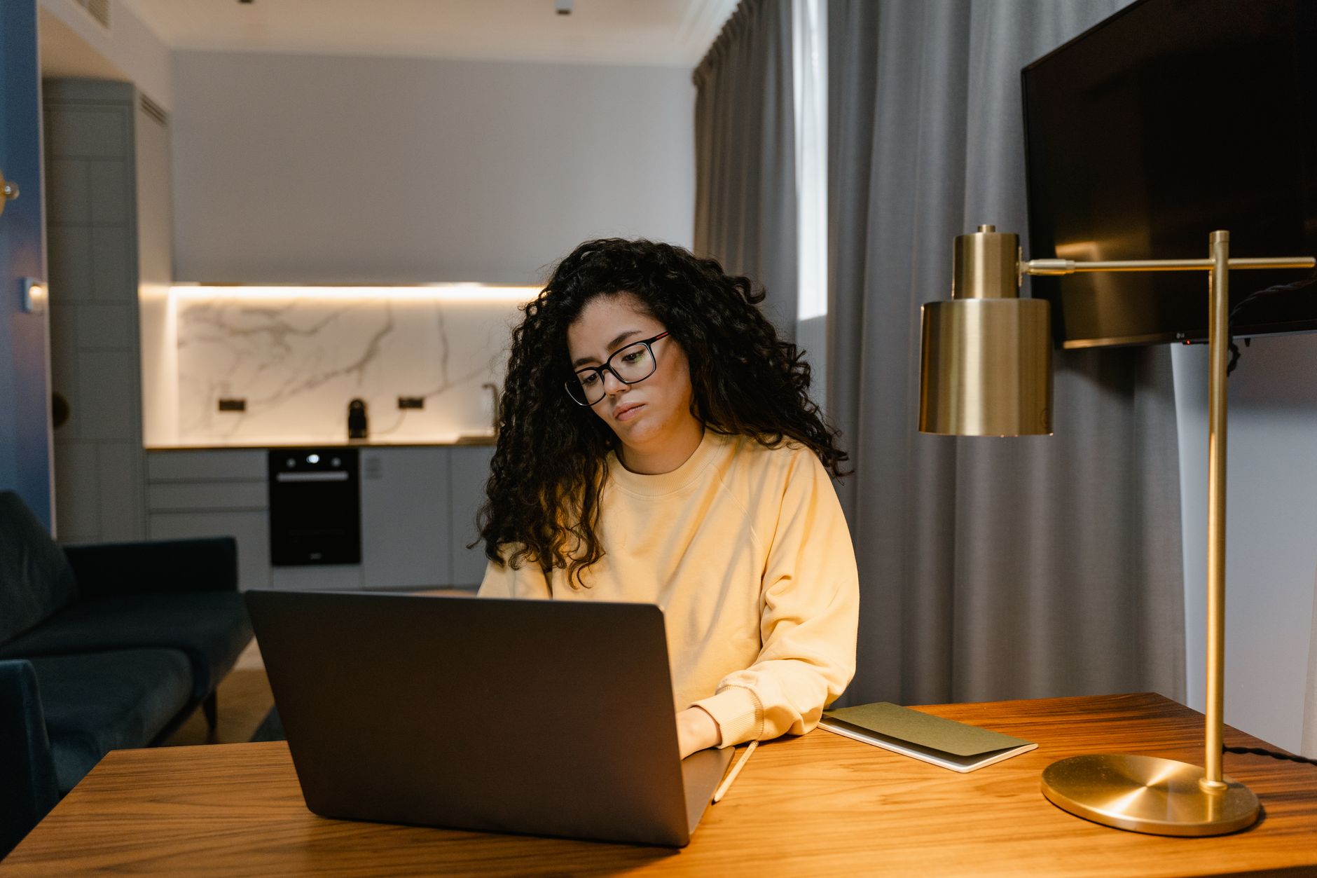Kvinde i gul sweatshirt arbejder på en bærbar computer hjemme i et hyggeligt, moderne rum.