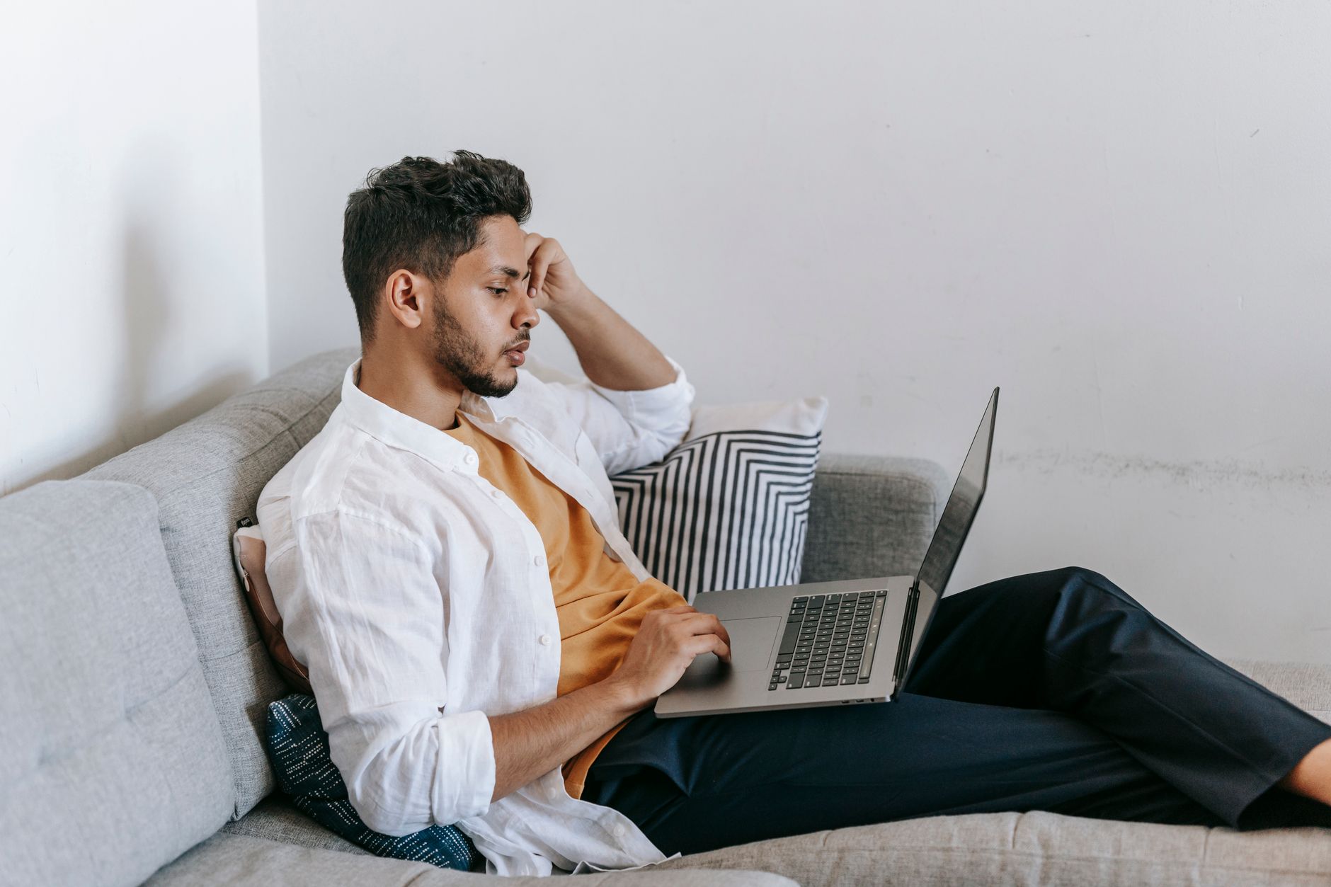 En ung mand ses fra siden, mens han sidder i en sofa og arbejder på en bærbar computer hjemmefra.